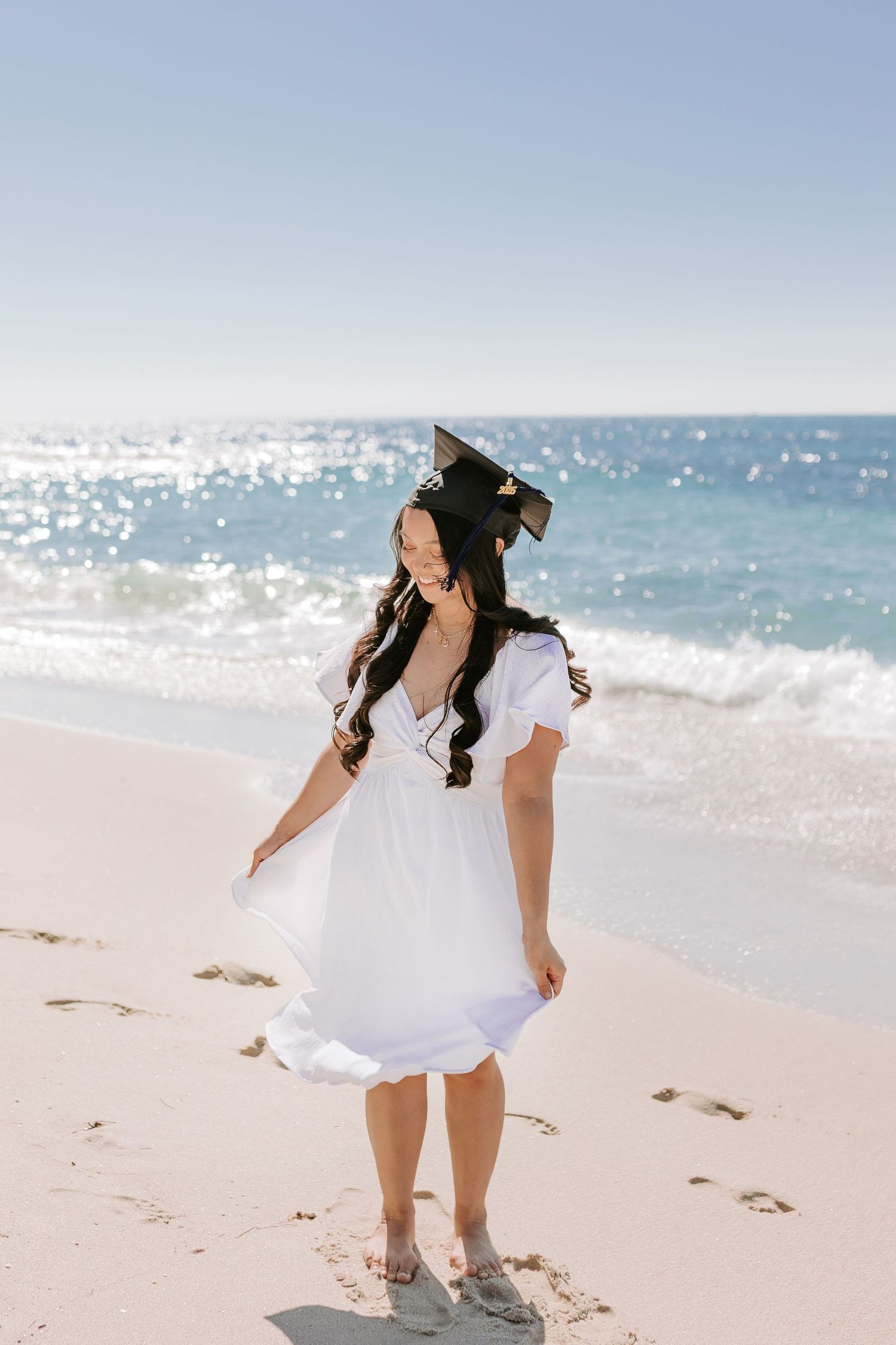 Graduate wearing a white dress smiles on the beach on a sunny day.