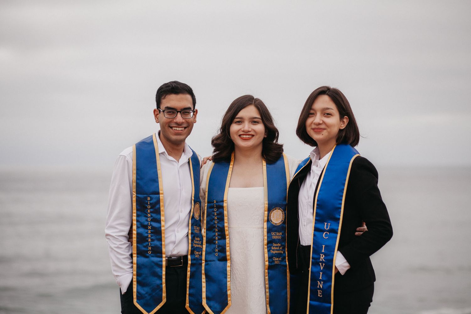 A group poses together wearing blue graduation sashes on a cloudy day with the ocean.