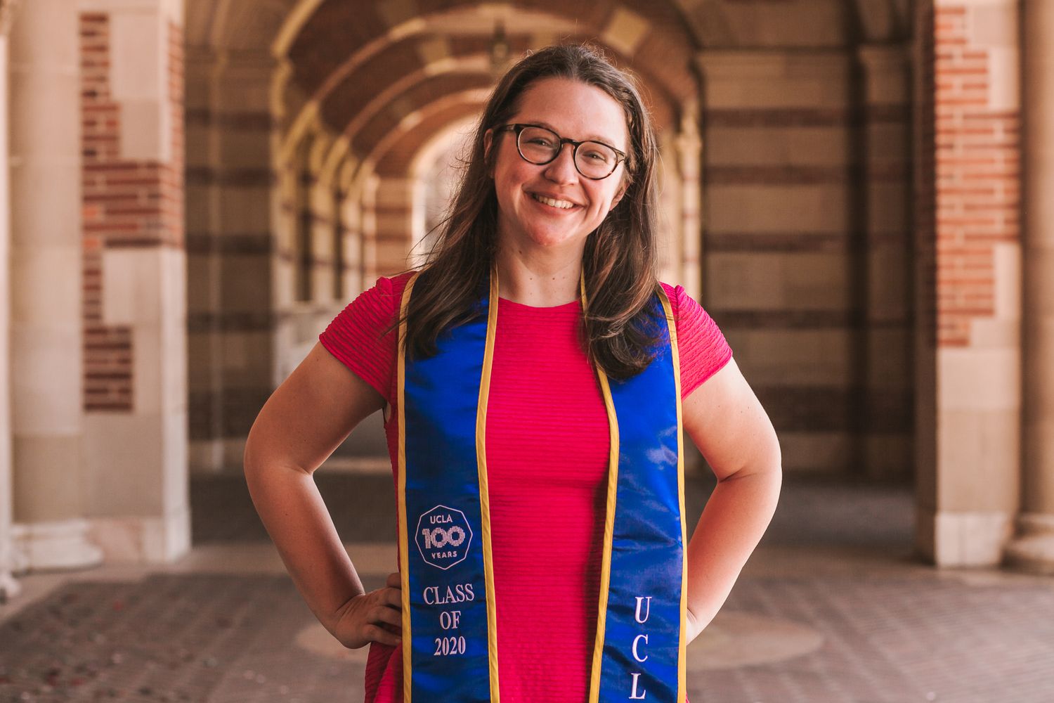 A woman wearing a pink dress and blue graduation sash stands in the halls of UCLA.