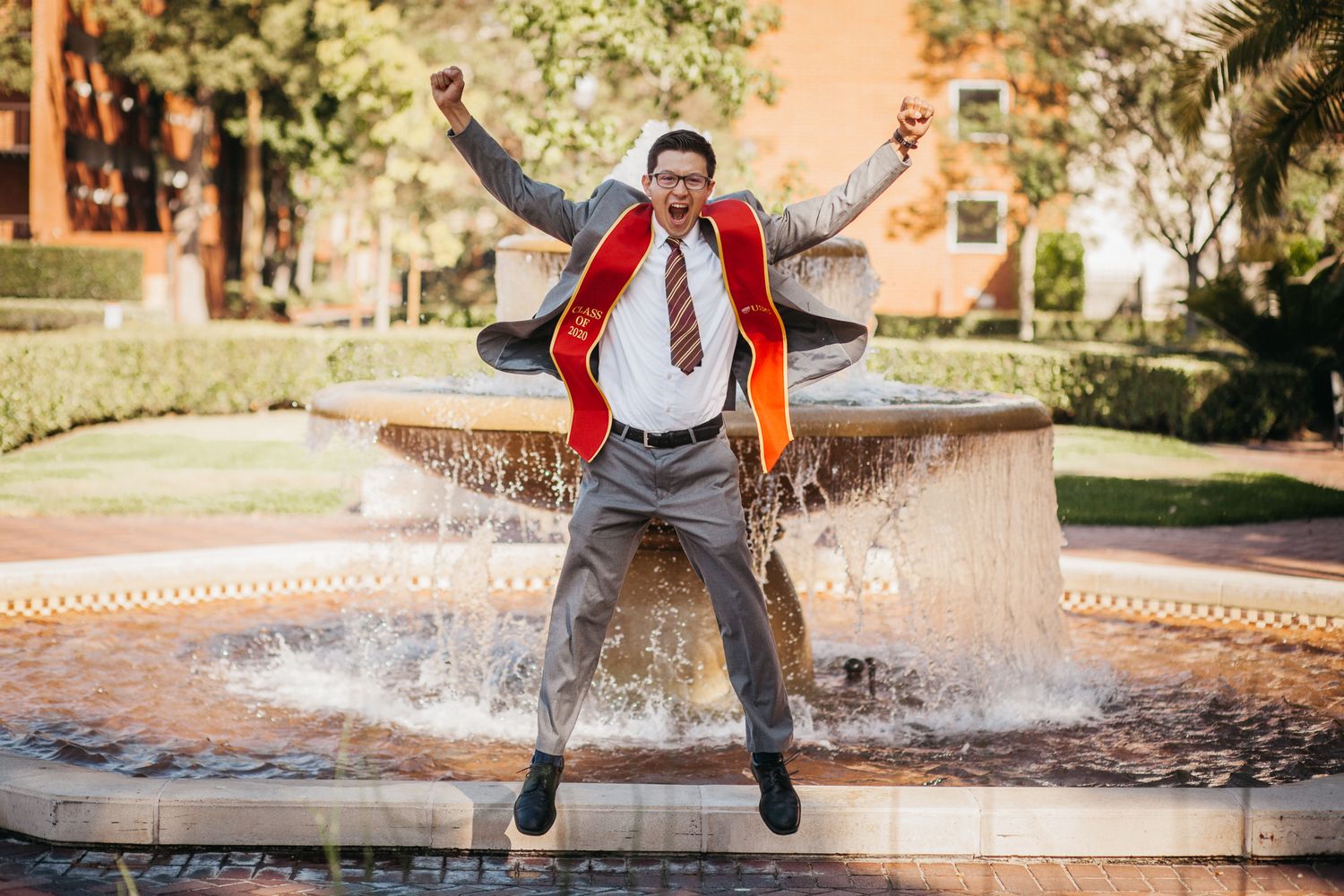 A man wearing a suit and graduation sash jumps in front of a fountain at USC.