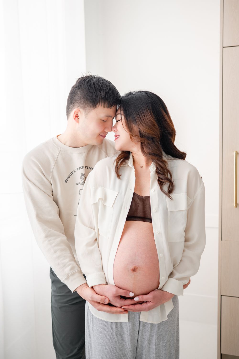 Intimate couple in white and brown attire share a tender moment while cradling a visible baby bump.