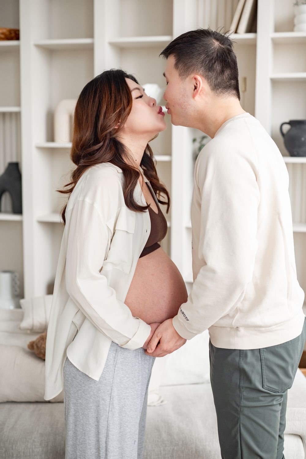 A romantic maternity moment shared between a couple in white sweaters against white built-in shelving.