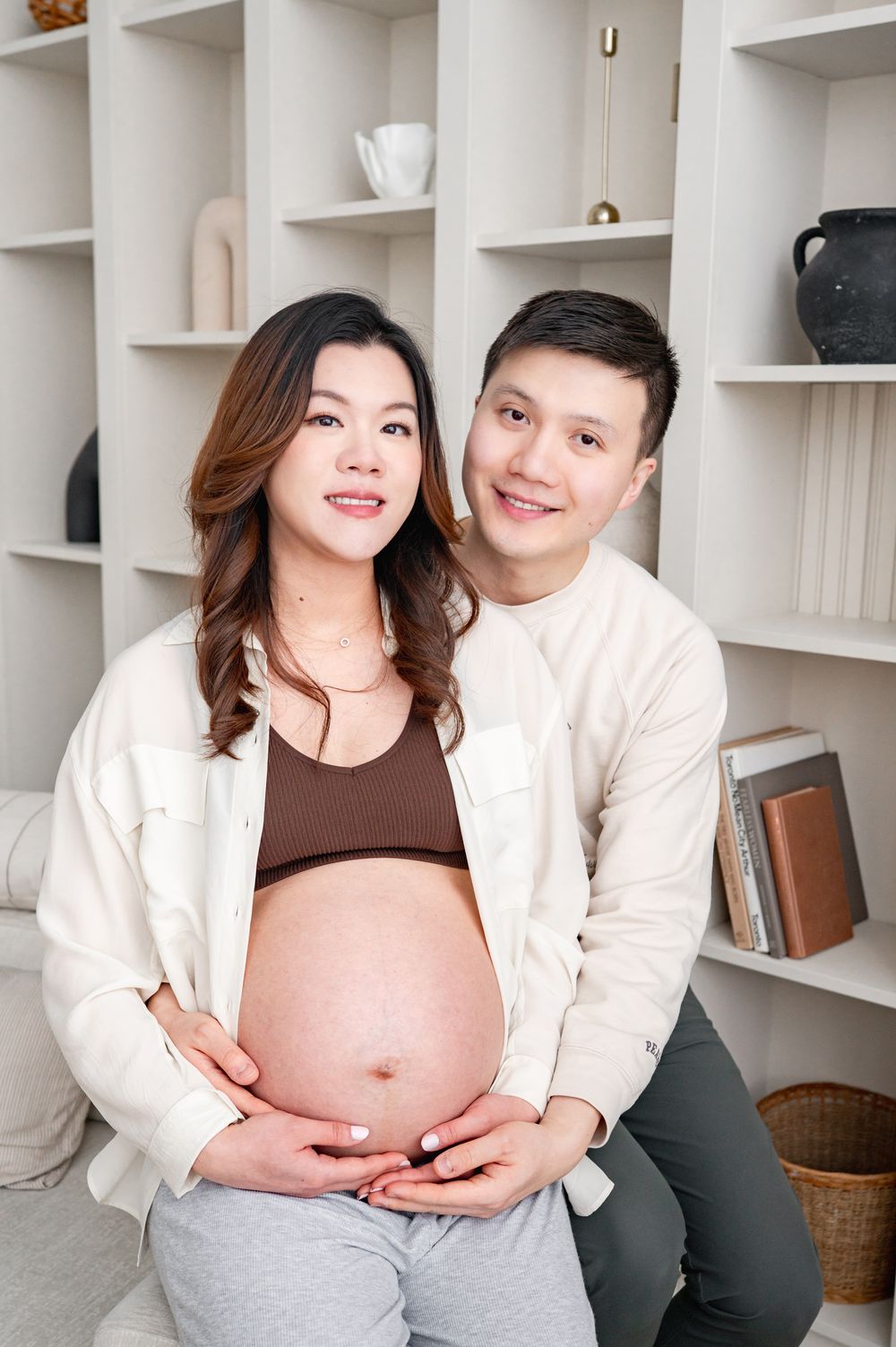 Expecting couple wearing matching cream cardigans pose together by white shelving unit with decorative items.