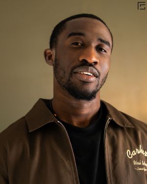 Person wearing a black t-shirt and brown jacket smiles warmly while posing against neutral background.