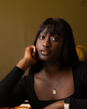Portrait photograph against a warm yellow background showing someone wearing a black top and delicate lock necklace.