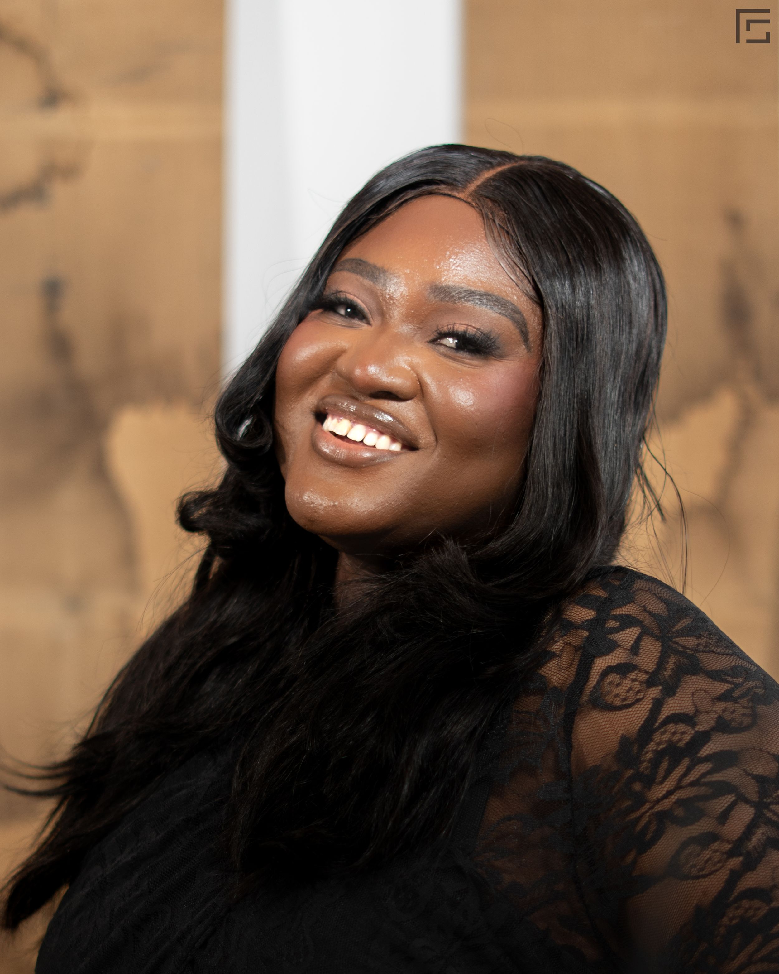 Portrait of someone with long black hair wearing lace sheer top, showing a bright smile against warm brown background.