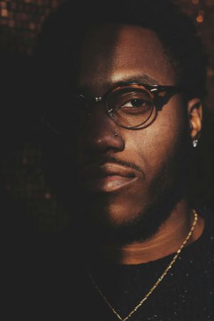 Artistic portrait of person wearing glasses and gold chain necklace in moody lighting against dark background.