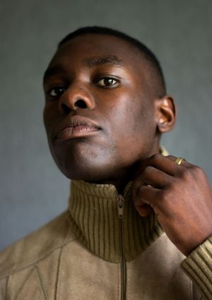Intimate portrait of person in beige turtleneck sweater touching their face against grey background.