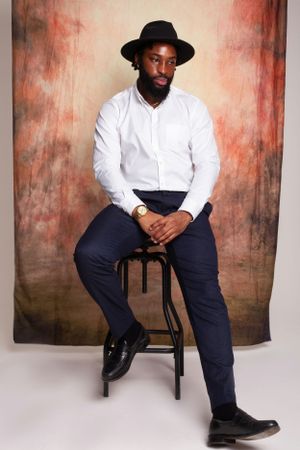 Stylish portrait of person in white shirt and black hat seated on stool against textured backdrop.