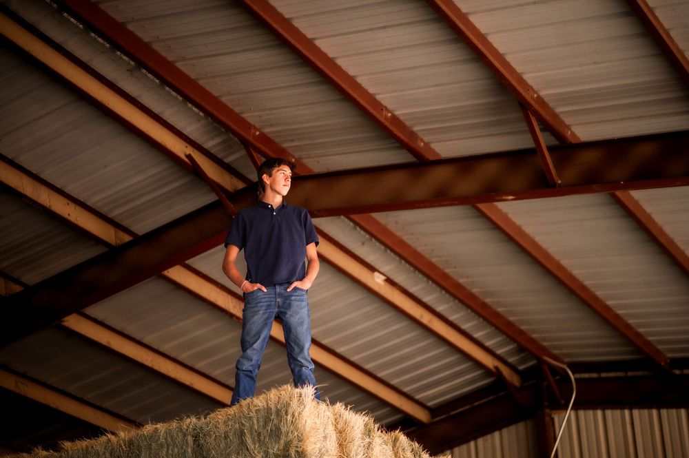 Man standing confidently on hay bales inside a rustic barn with metal roof and wooden beams.