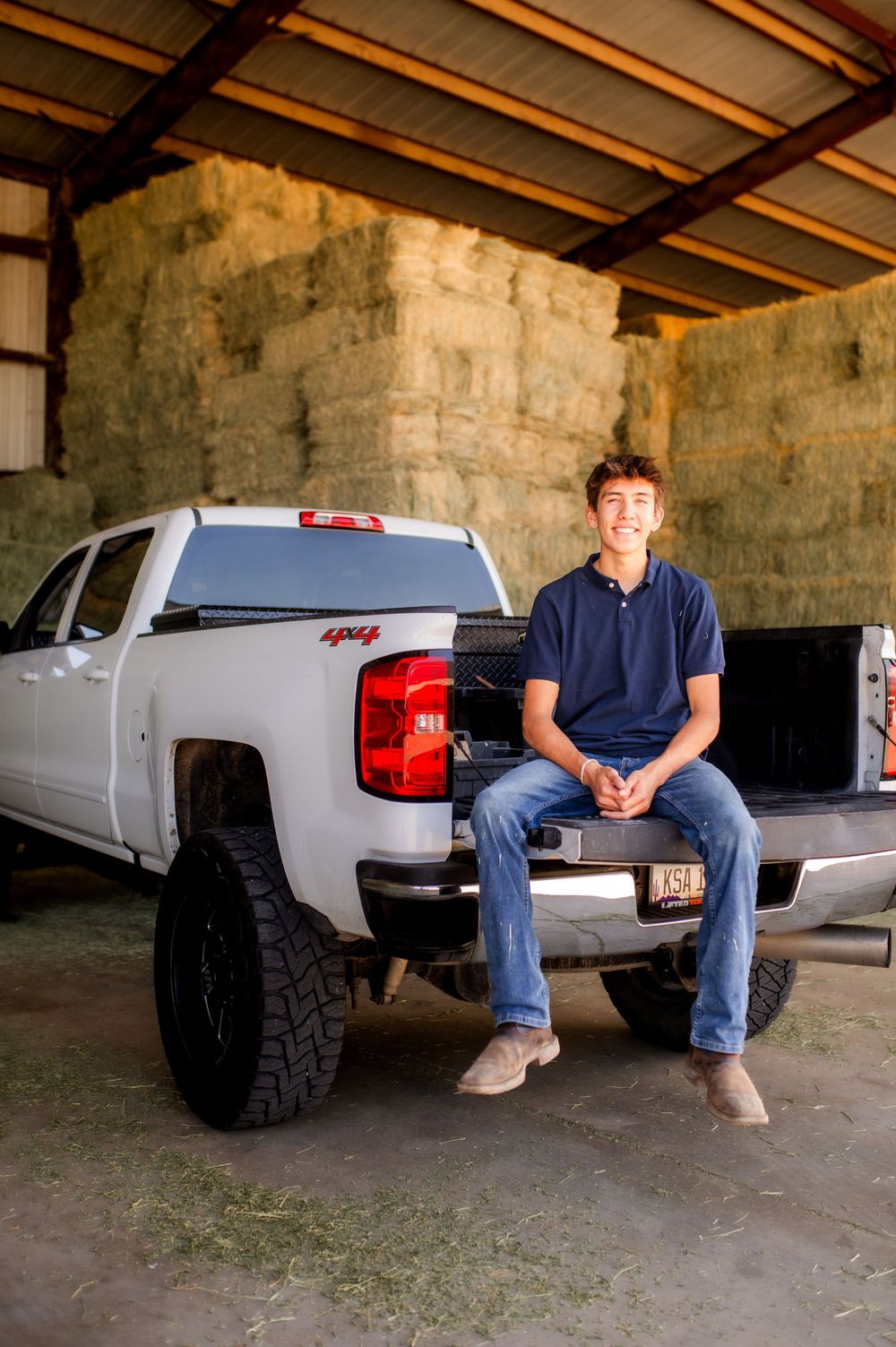 Young man sitting on tailgate of white Chevy 4x4 truck in hay barn, smiling casually in jeans and navy polo.