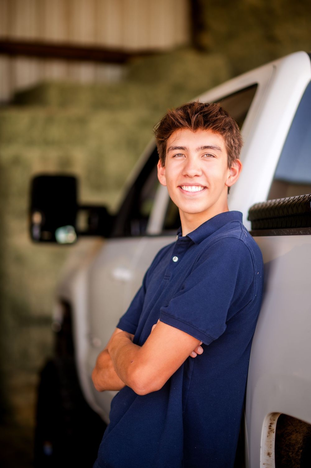 Smiling young man in navy polo shirt leaning against white truck with arms crossed in a barn setting.