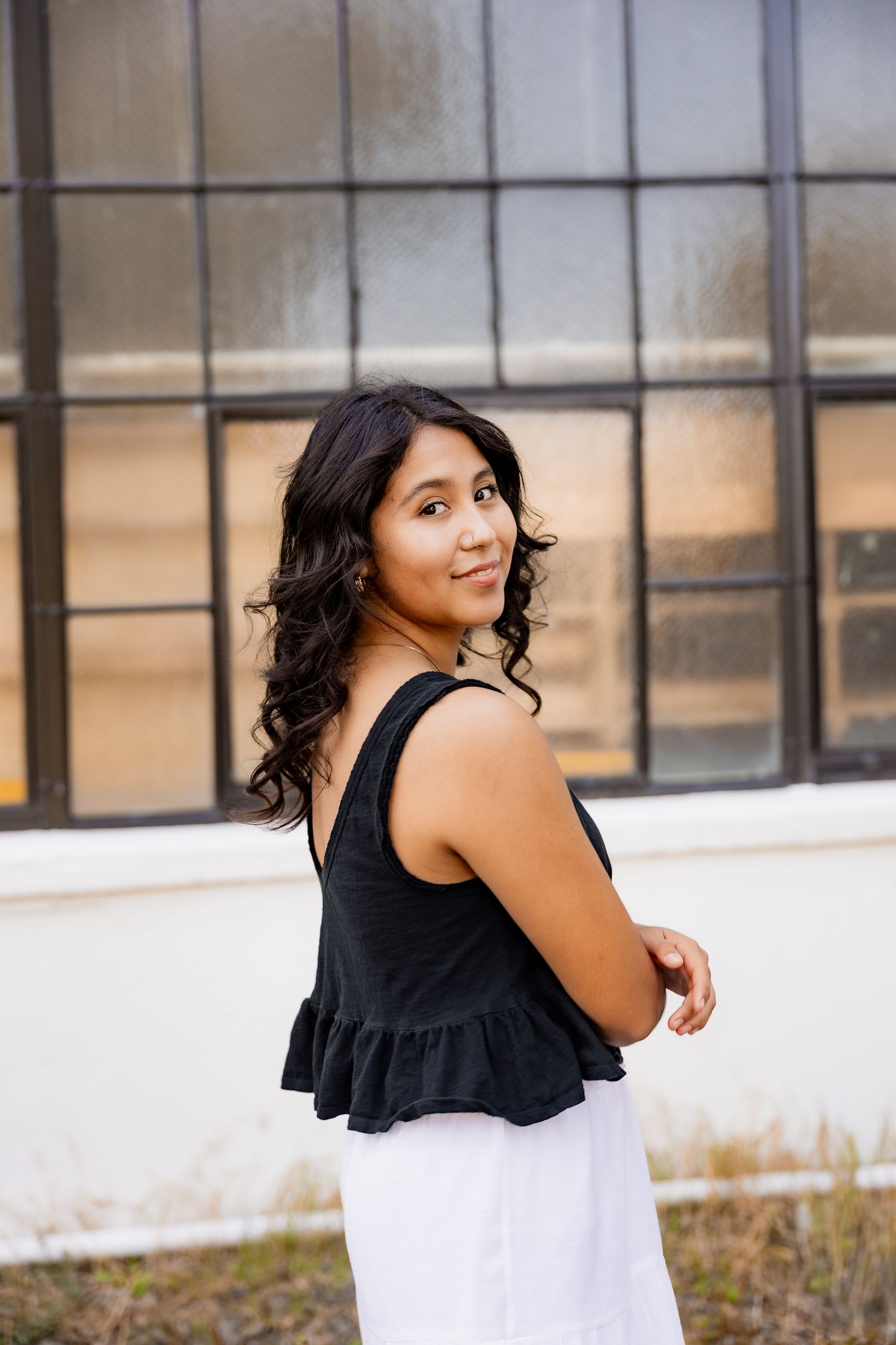 A person in a black sleeveless top and white skirt poses outdoors against an industrial building with windows.