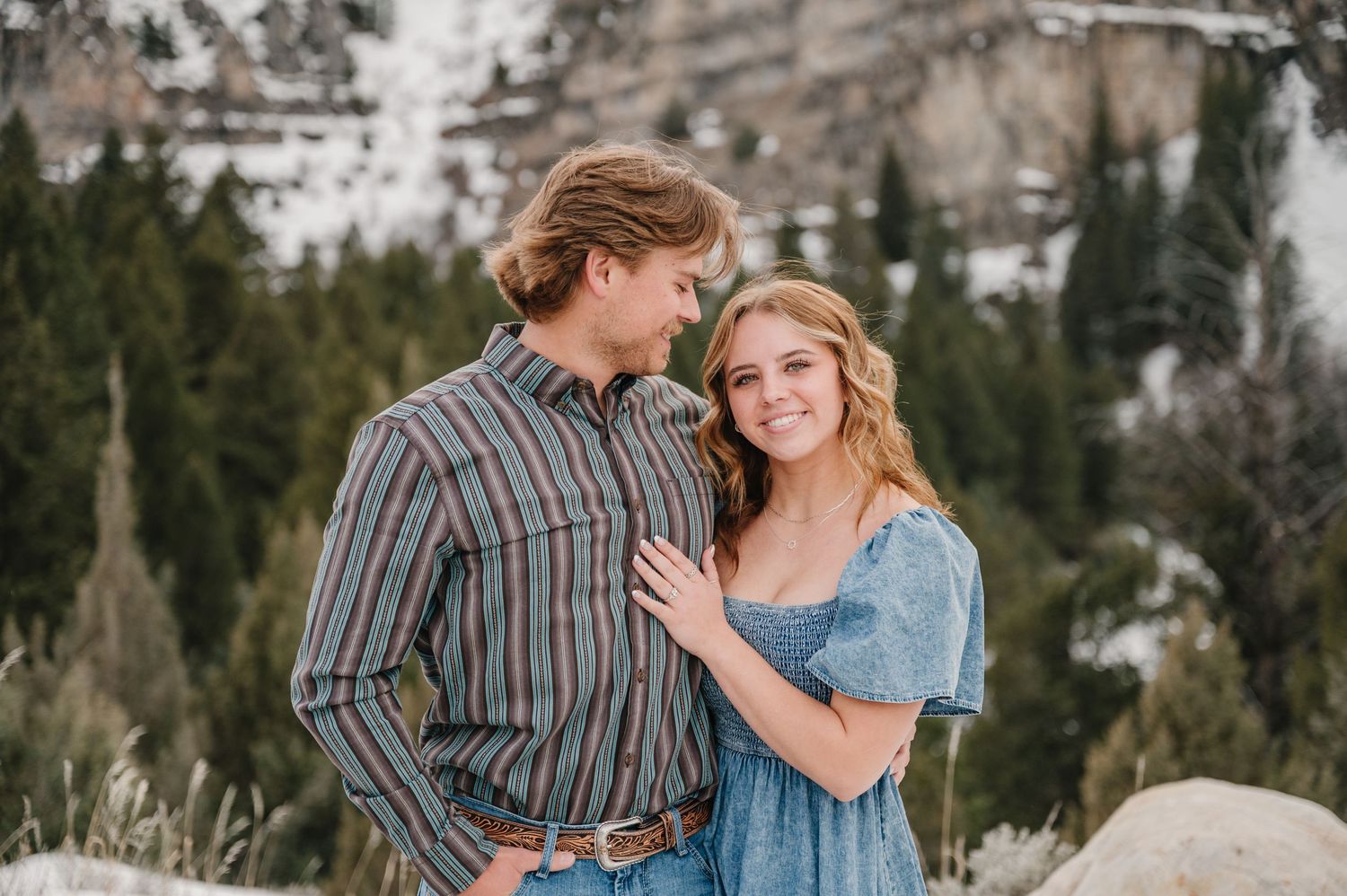 A romantic couple poses together in front of a snowy mountain landscape wearing casual western attire.