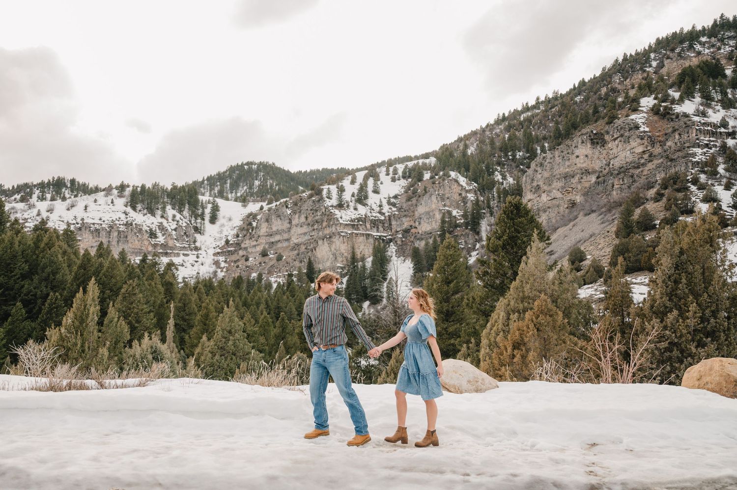 A couple shares a romantic moment in the snow with snowy mountains and a waterfall in the backdrop.