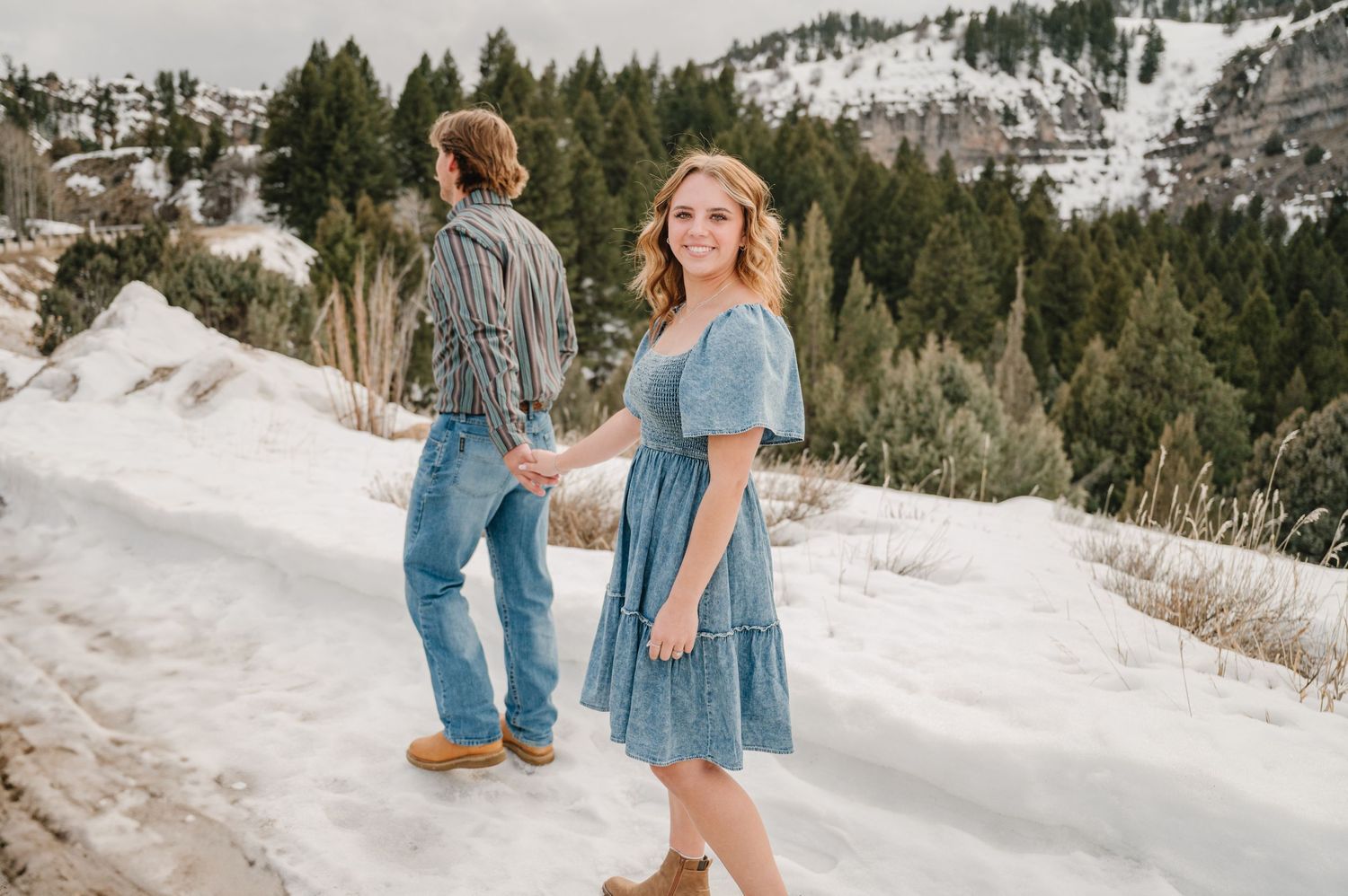 A couple strolls through a snowy mountain landscape with evergreen trees and snow-covered slopes in the background.