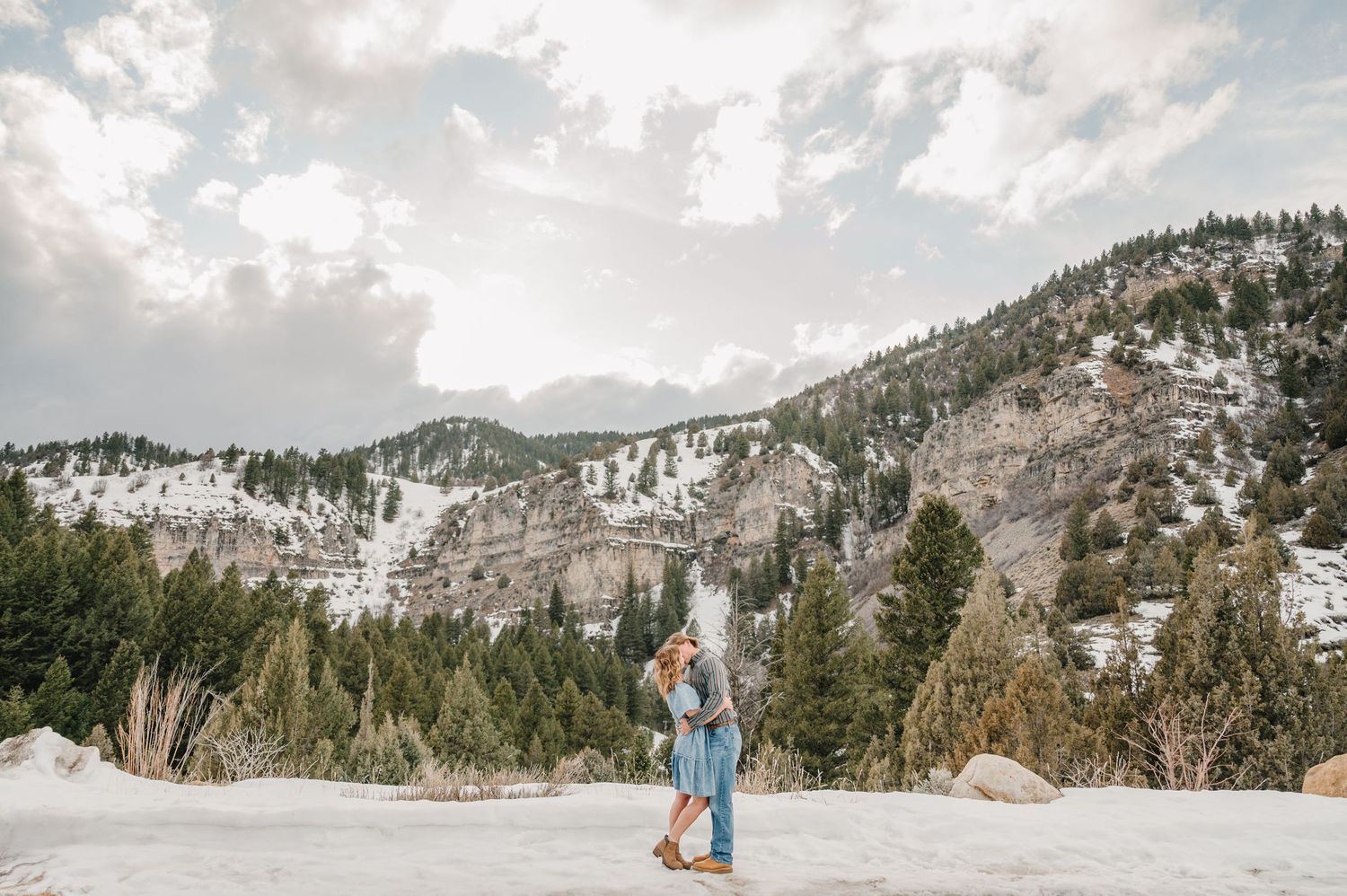 A couple embraces in a snowy mountain landscape with evergreen trees and snow-capped peaks in the background.