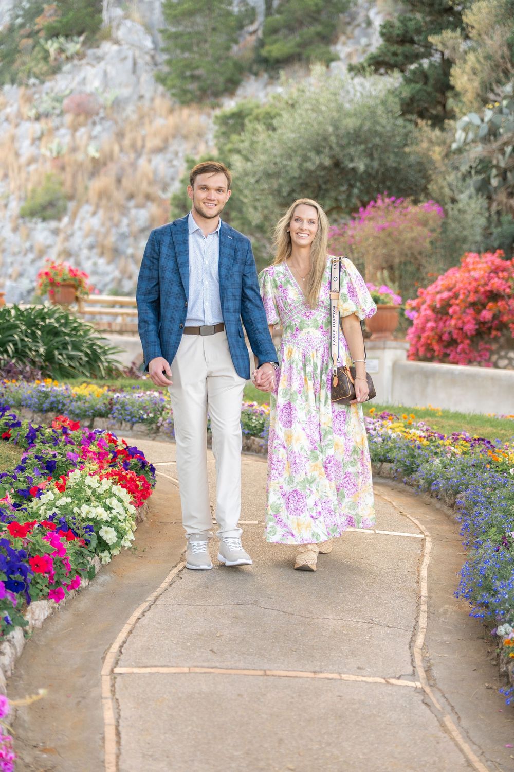 A couple walks through a vibrant garden path lined with colorful flowers and blooming azaleas on a sunny day.