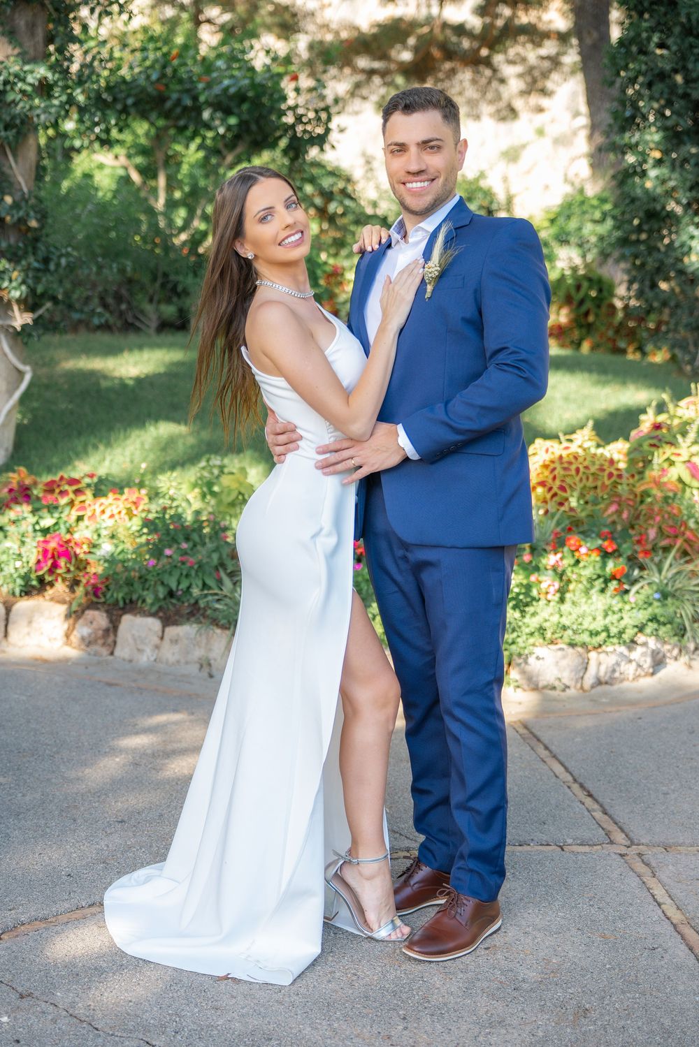 A couple poses together in a garden setting, wearing a white gown and blue suit on their wedding day.