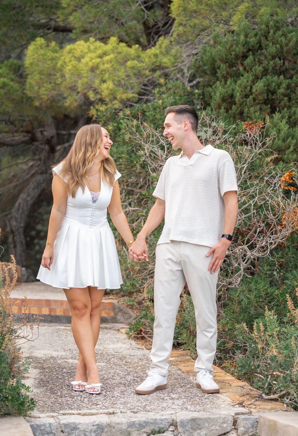 A couple wearing white outfits walks together on a stone path surrounded by greenery and flowers.