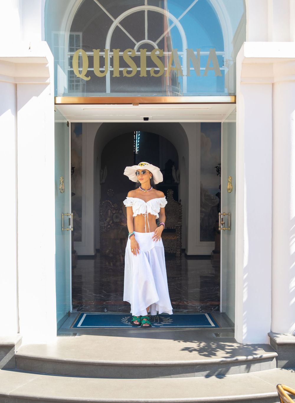 Someone in a white summer outfit with a wide-brimmed hat stands in an elegant doorway beneath a curved window.