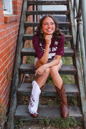 Smiling young woman in Texas A&M shirt sits on metal stairs wearing cowboy boots.