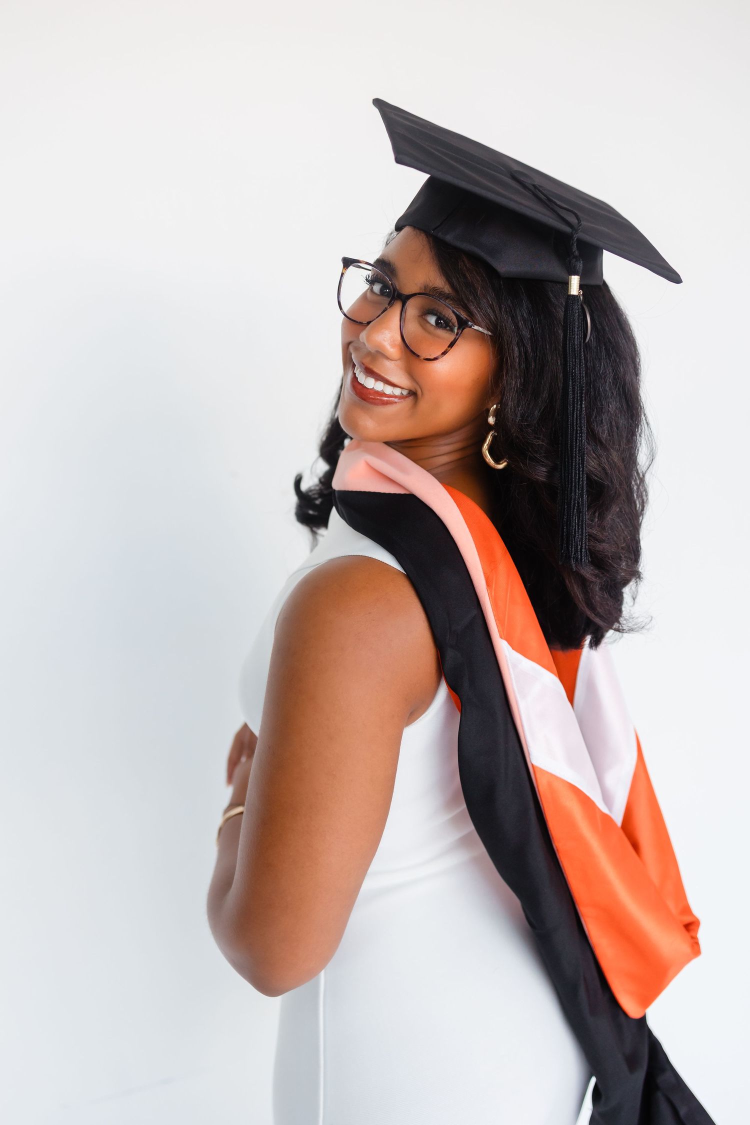 Smiling young Black woman in graduation cap and glasses with orange and white sash against white background.