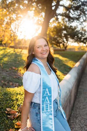Brunette woman in white top wearing light blue Alpha Delta Pi sorority graduation stole in golden sunset park.