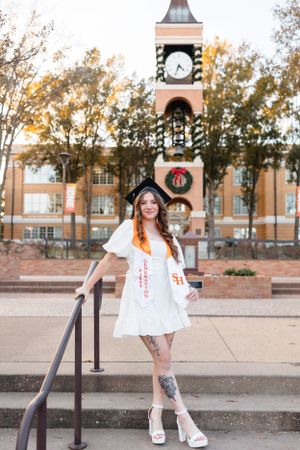 Young woman in white dress and graduation cap posing on steps before brick clock tower with holiday wreath.
