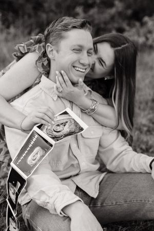 Happy couple sits in grass holding ultrasound photos in black and white maternity announcement photo.
