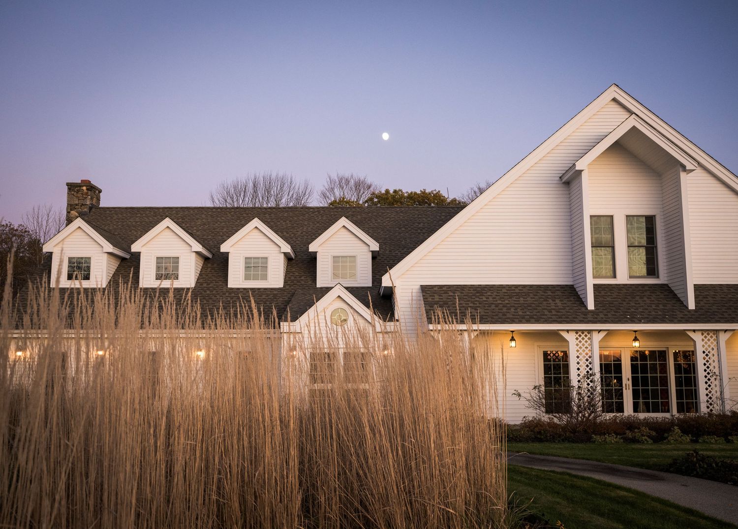 A white Cape Cod style house with green shutters features a wraparound porch and two dormer windows under a blue sky.