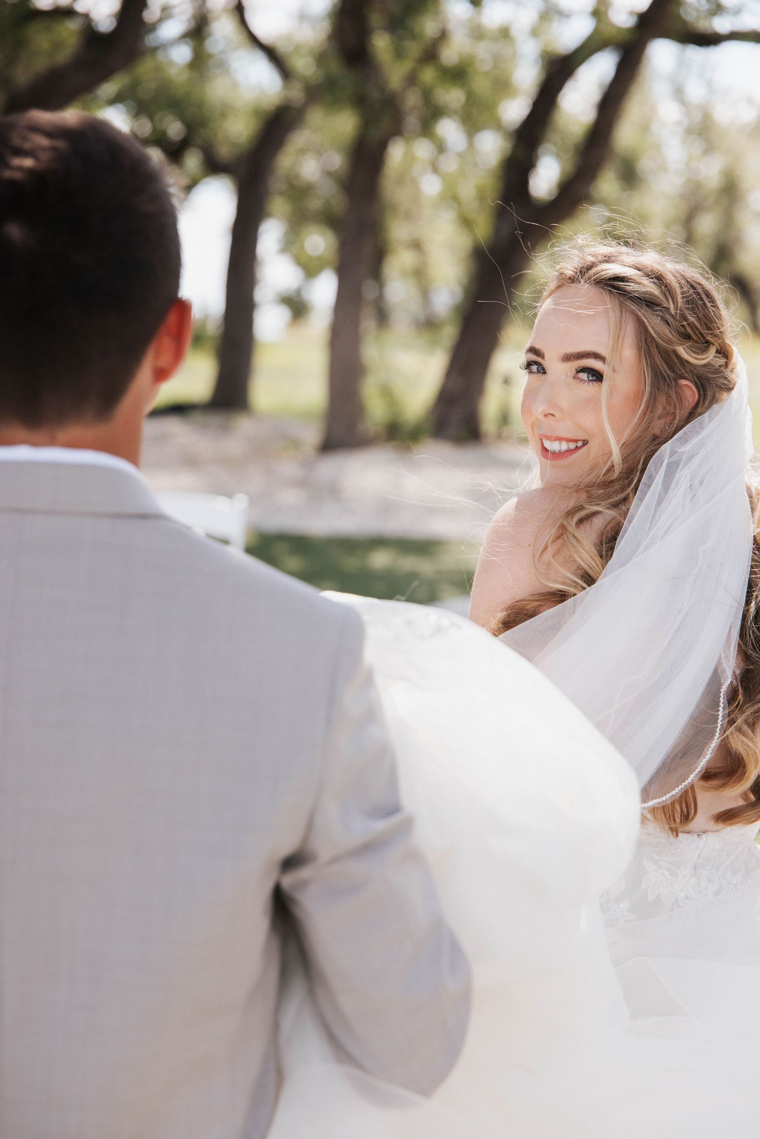 A bride wearing a white veil smiles while looking over her shoulder in a sun-dappled outdoor setting.