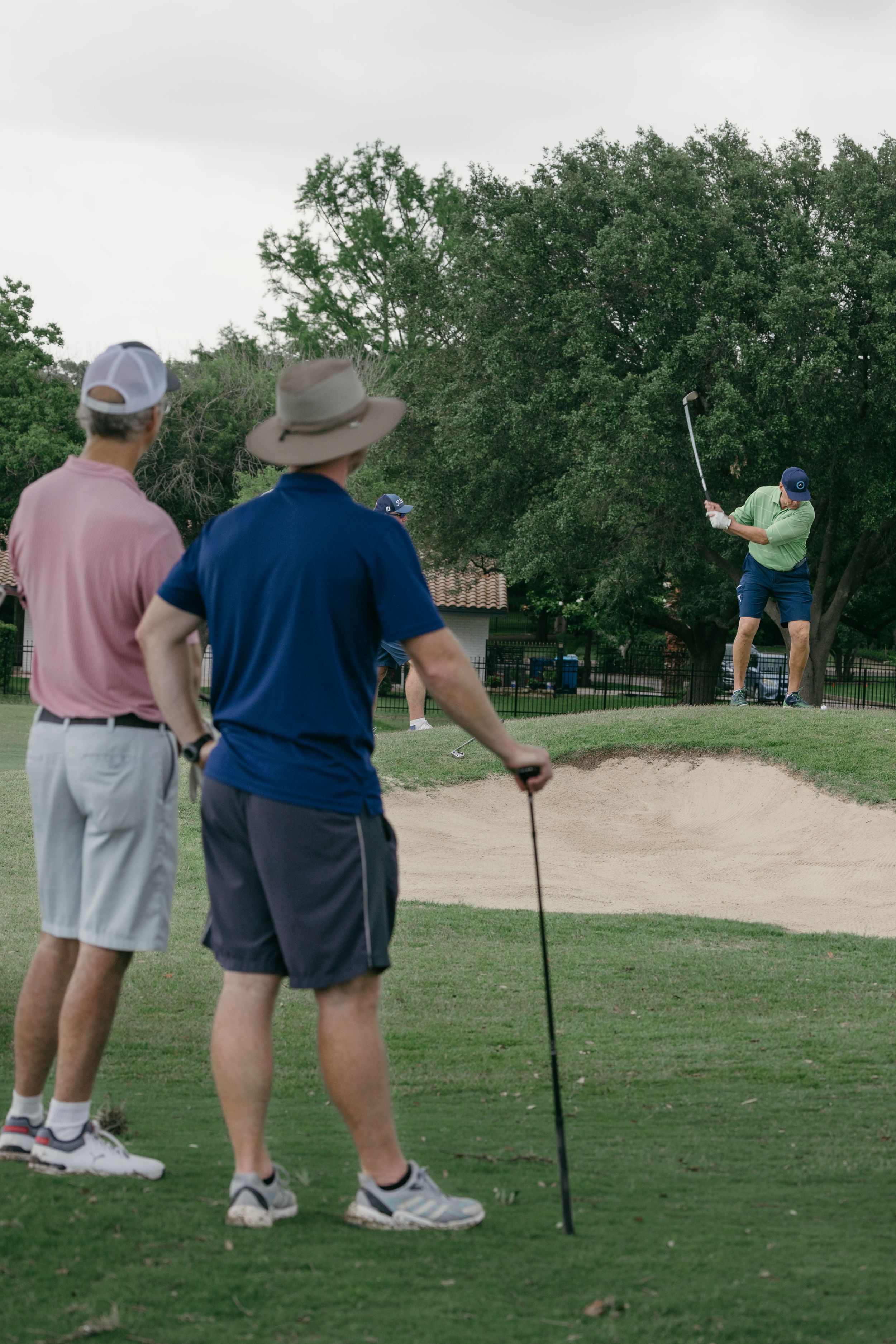 Golfers watching a player hit from a sand bunker on a lush green golf course with trees in the background.