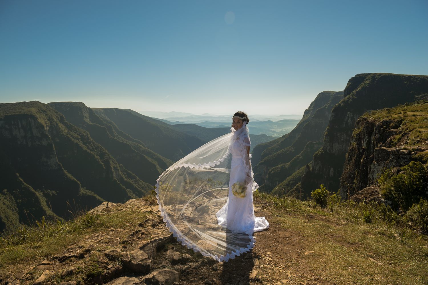 Bride in flowing dress stands on mountain cliff with scenic valley view.