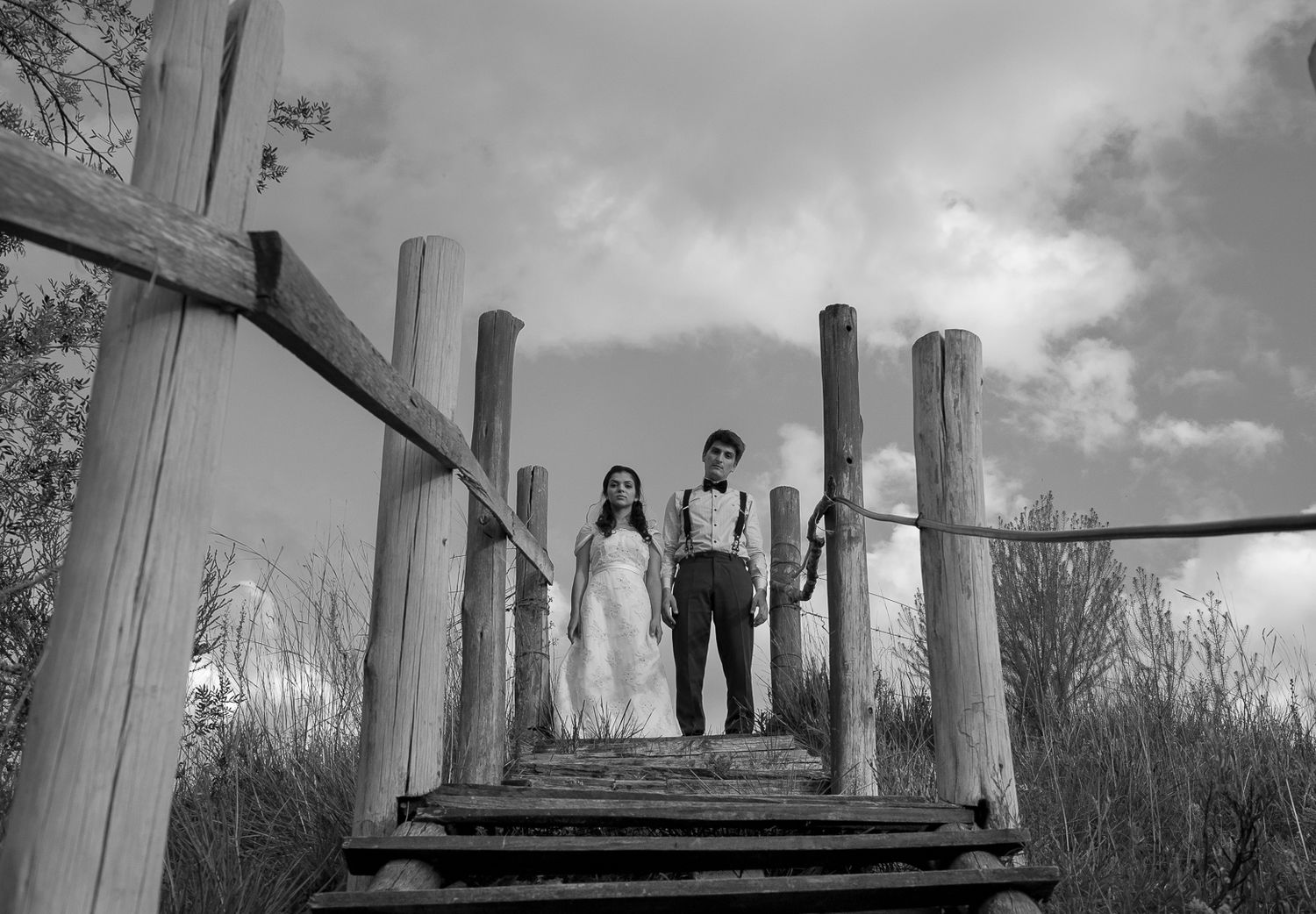 A couple stands together on a rustic wooden bridge against a cloudy sky.