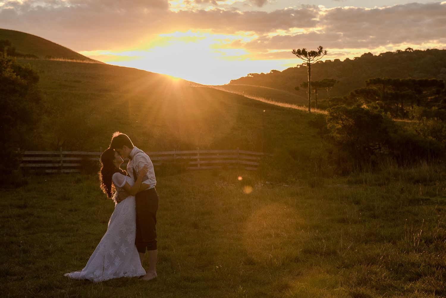 A couple embraces during golden hour sunset with rolling hills in the background.