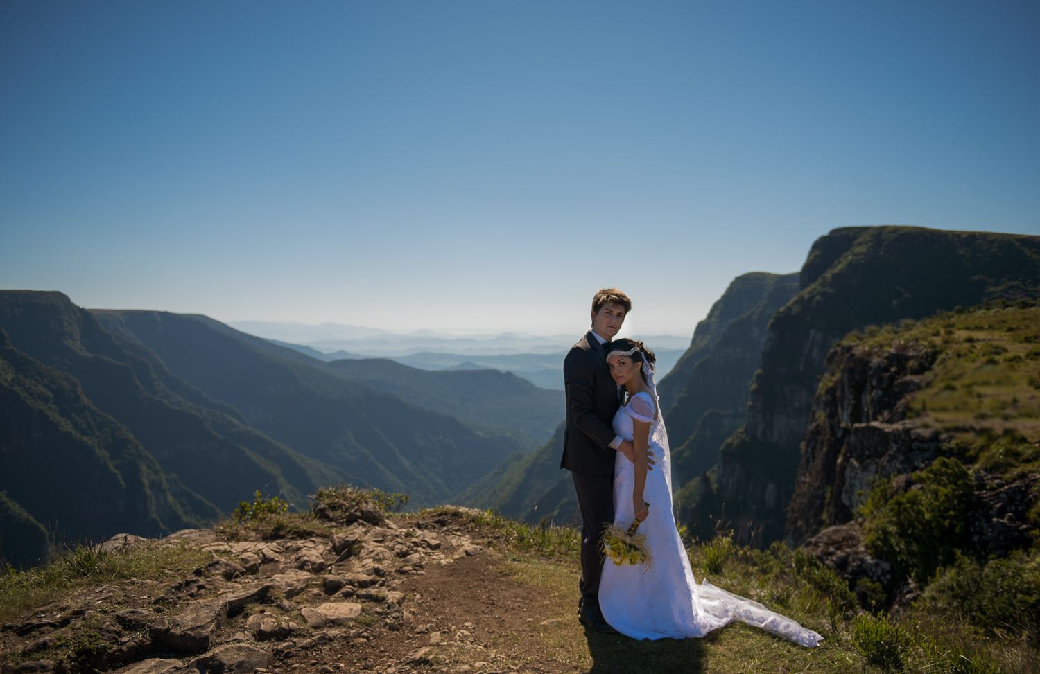 Wedding couple embracing on mountain cliff with panoramic valley view.