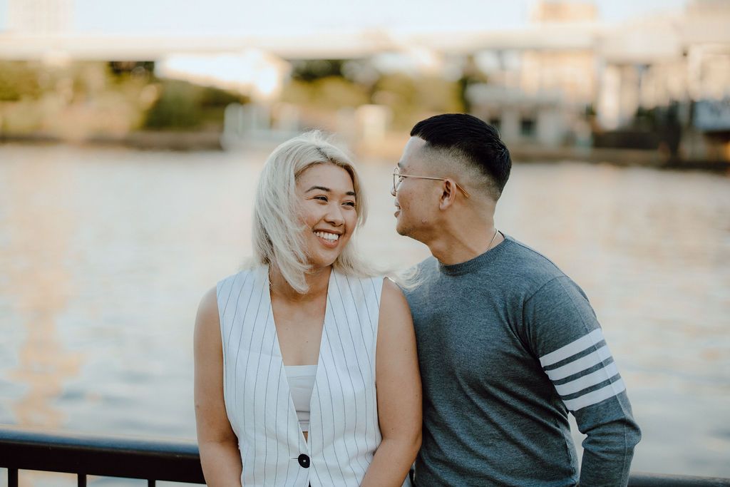 Couple sharing a romantic moment while standing by a riverside with a bridge in the background.