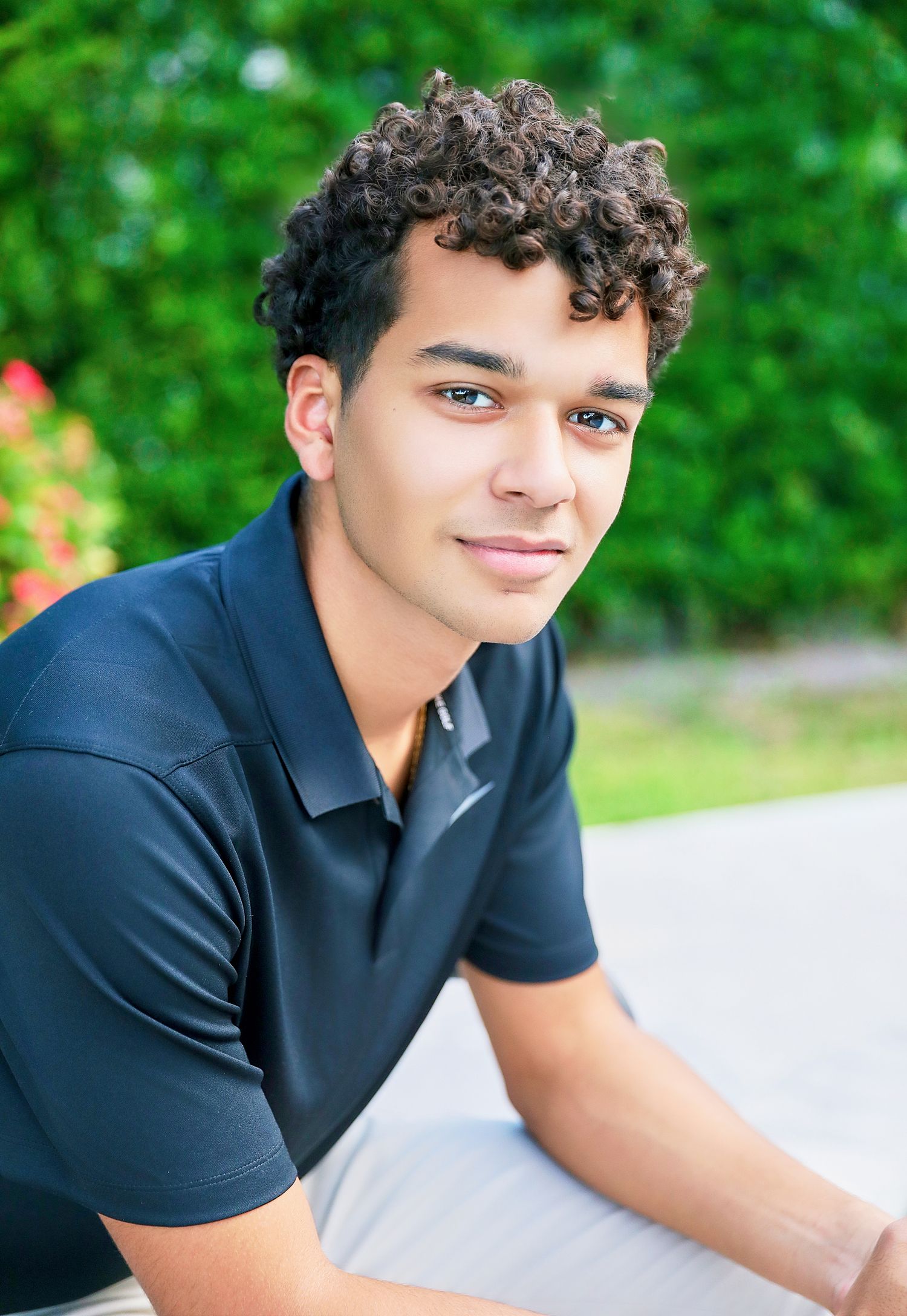 Young man in navy polo shirt smiles outdoors with curly hair.