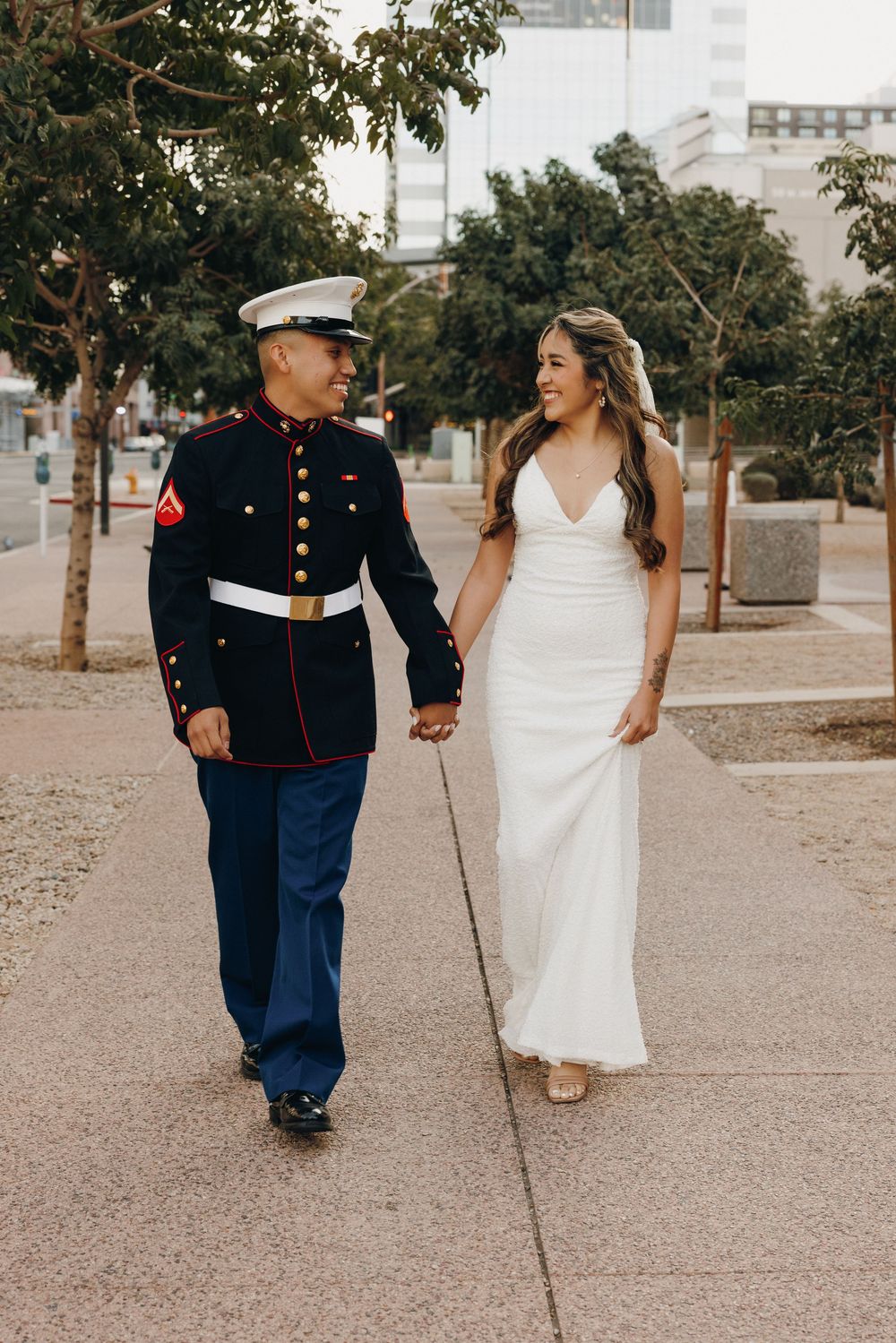 A bride in a flowing white dress walks alongside a Marine in dress uniform on a paved path outdoors.
