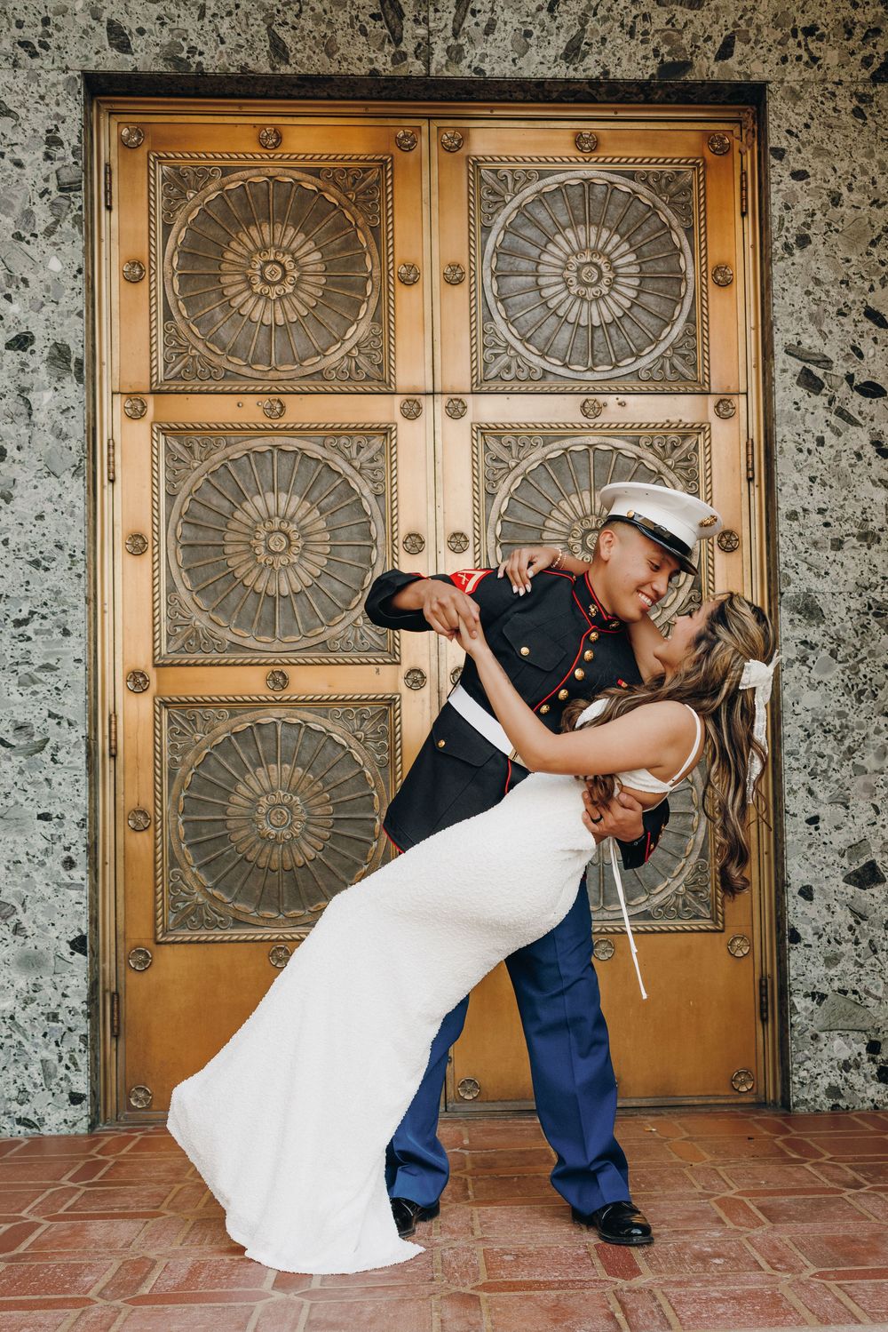 A couple in wedding attire pose in front of ornate bronze doors with decorative sunburst panels.