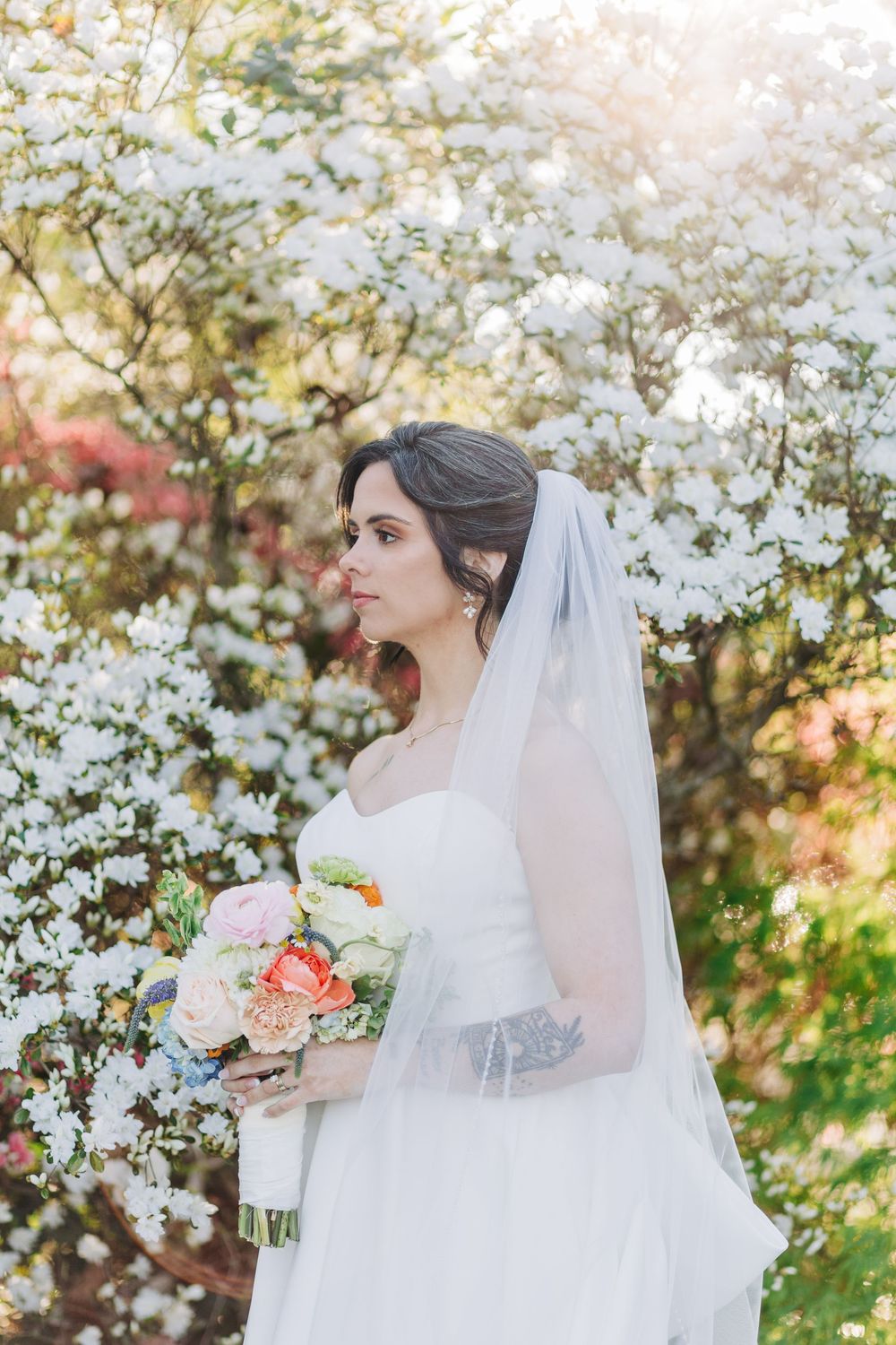 Bride in white gown and veil holds colorful bouquet among blooming white flowering trees in soft sunlight.