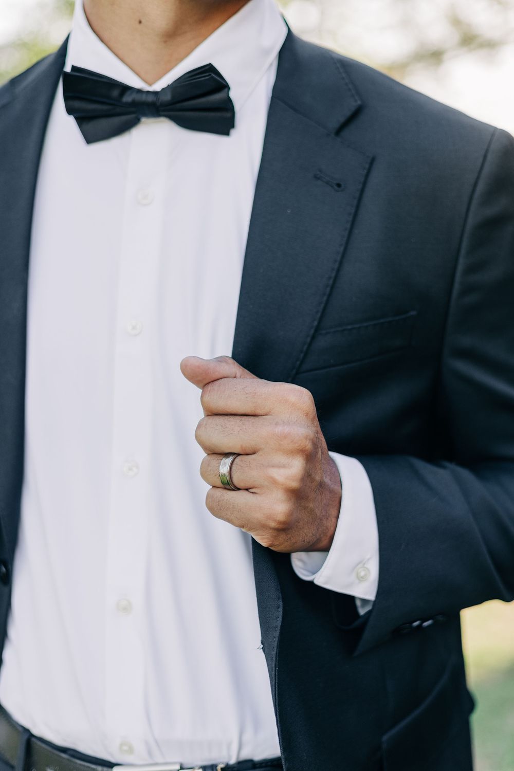 Man in dark suit with black bow tie and wedding ring holding lapel, elegant groom attire for formal occasion.