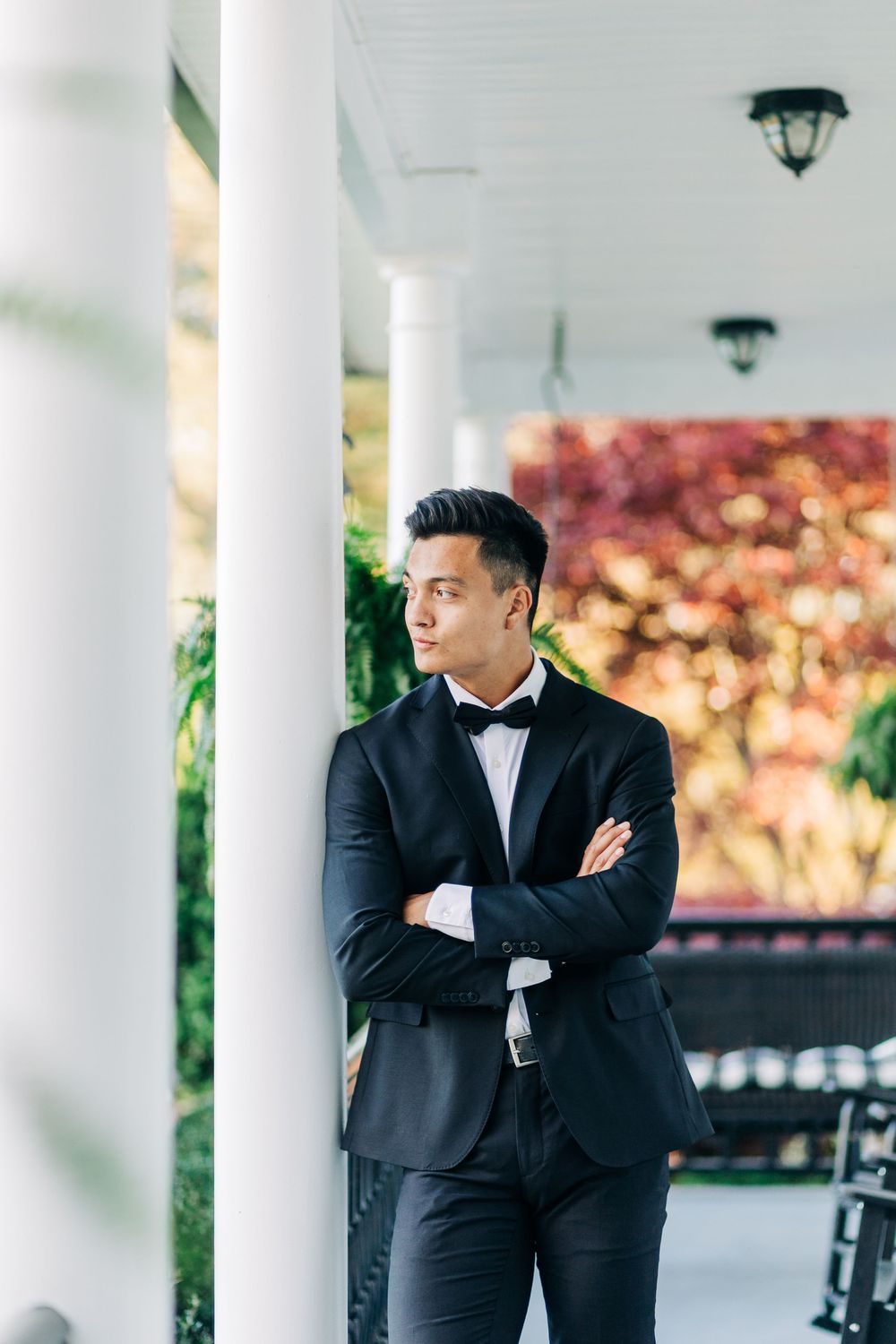 A young man in a black tuxedo with bow tie stands confidently with arms crossed beside white columns on an elegant porch.