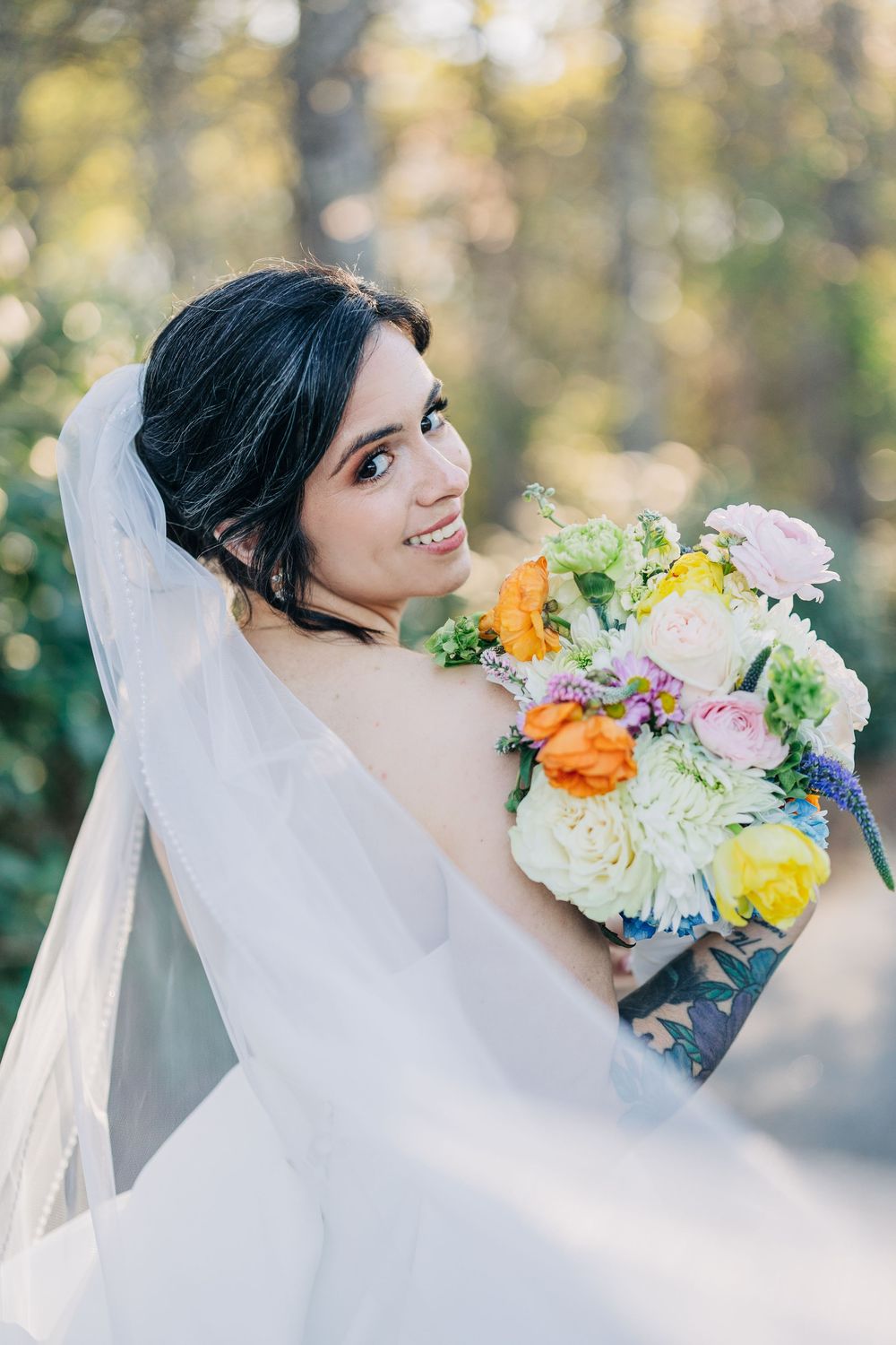Smiling bride in white gown and veil holds colorful bouquet with orange, pink, and white flowers outdoors.
