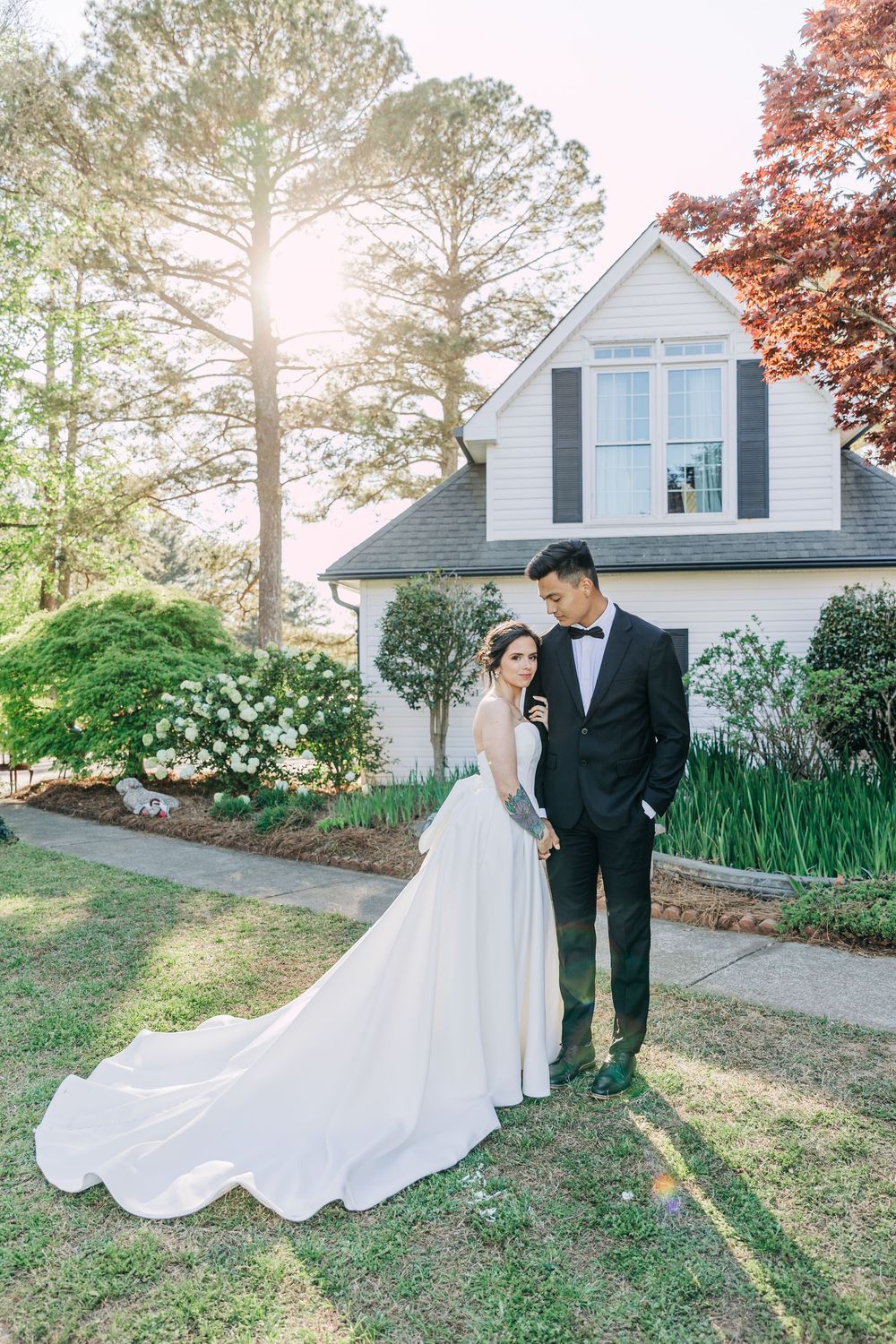 Bride and groom stand together in front of a white house surrounded by lush greenery on their wedding day.