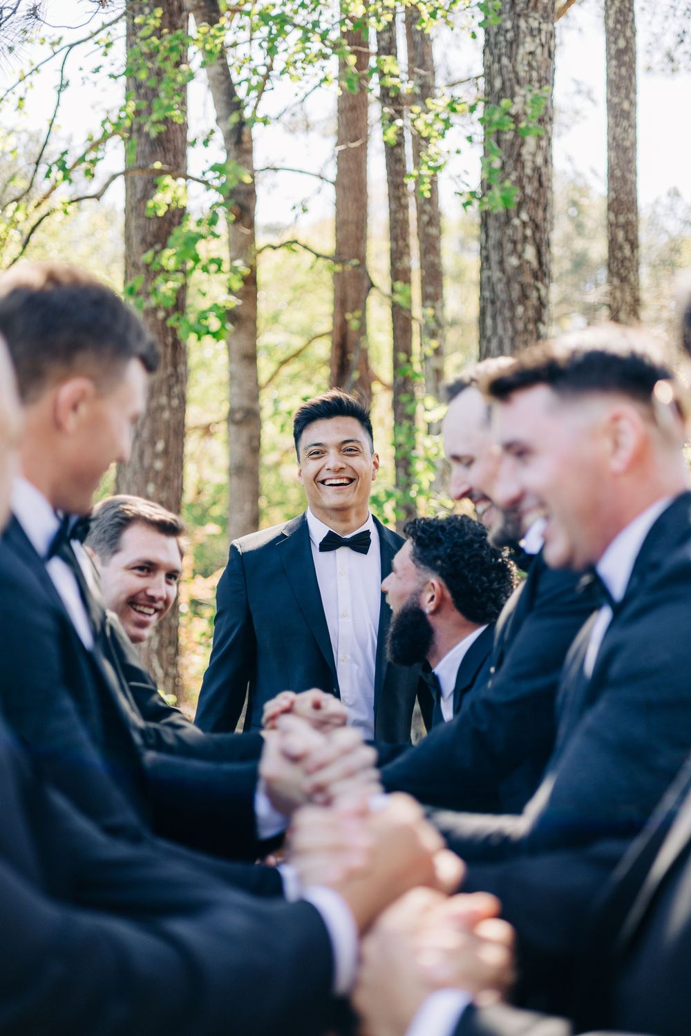 Groom laughing with groomsmen in suits and bow ties, celebrating outdoors in a sunlit forest setting.
