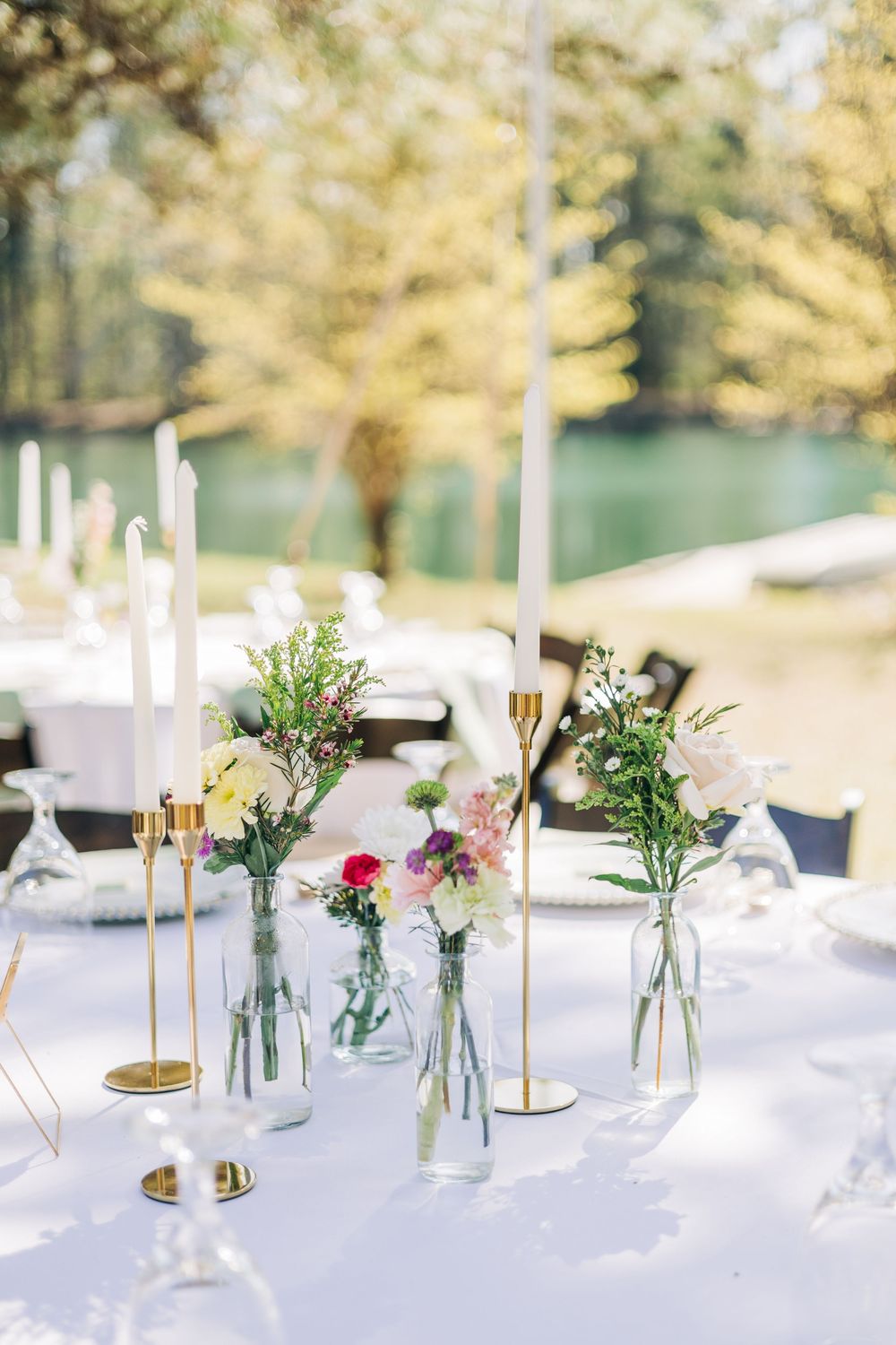Elegant outdoor wedding table with white linen, gold candlesticks, and colorful wildflowers in glass bud vases.