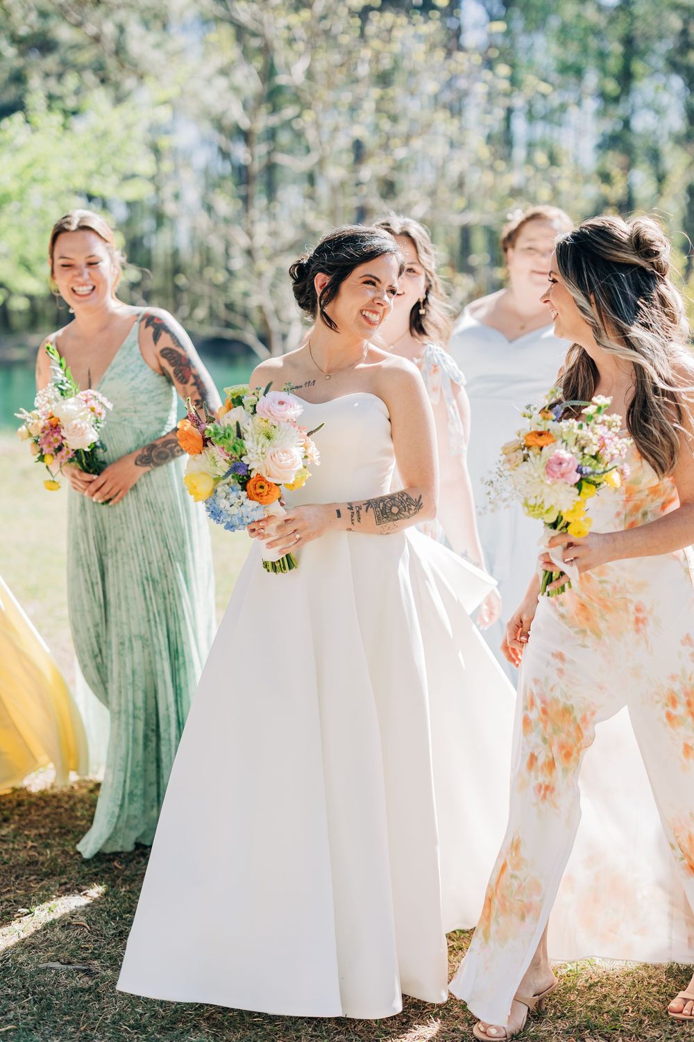 Bride in white strapless gown walks with bridesmaids in colorful dresses, all holding floral bouquets outdoors.
