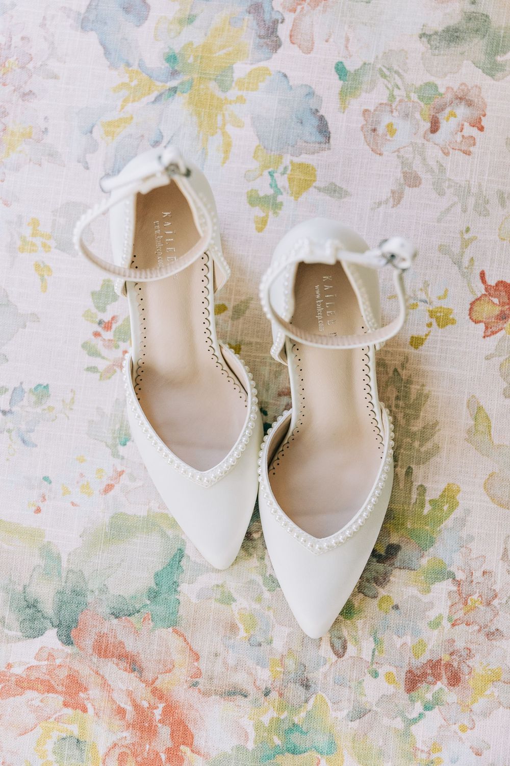 Elegant white pointed-toe bridal heels with ankle straps and bow details on a soft floral background.